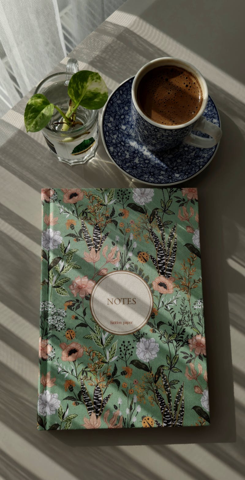 A quiet desk with an open notebook, a small ceramic mug, a sprig of greenery, and warm window light — a moment of stillness.